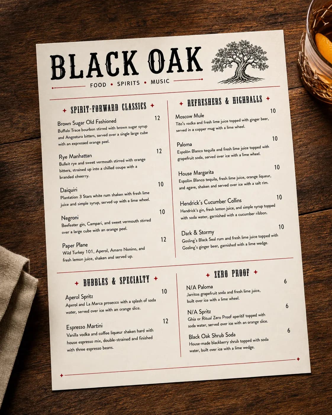 Black Oak cocktail menu photographed on a dark wood table beside a rocks glass, printed on cream paper with a black slab-serif 'BLACK OAK' wordmark, a 'food, spirits, music' rule, an oak tree engraving, and red rules dividing four cocktail sections.